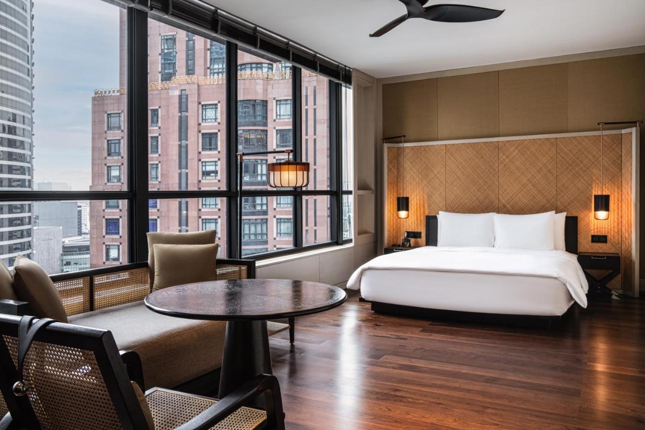 The Ruma Hotel And Residences Kuala Lumpur Exterior photo The photo shows a modern hotel room featuring a large bed with white linens, situated against a backdrop of large windows that allow for ample natural light. The room includes a wooden floor and has a minimalist aesthetic, with a round table and two 