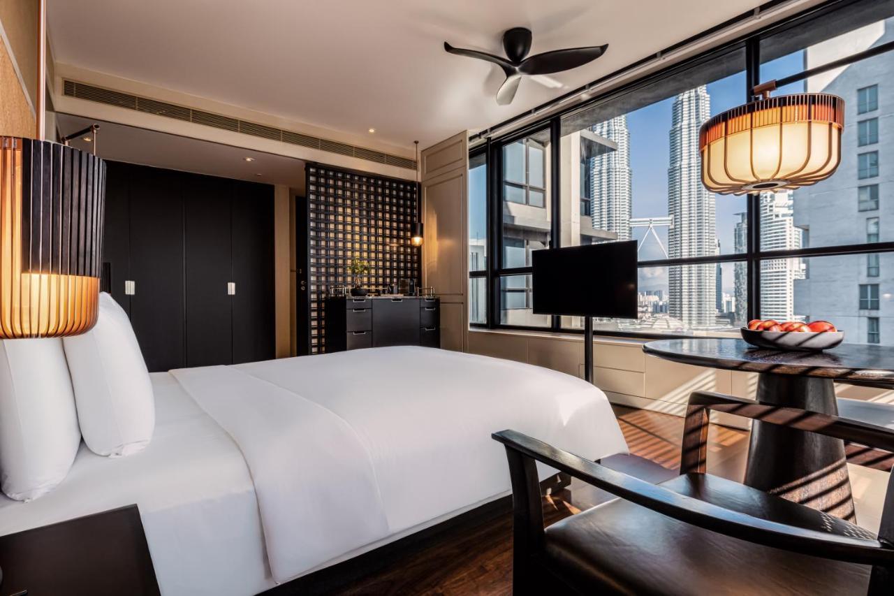 The Ruma Hotel And Residences Kuala Lumpur Exterior photo The photo shows a modern hotel room featuring a large bed with white linens. There's a ceiling fan above and large windows letting in natural light, providing a view of a cityscape outside. To one side, there's a small seating area with a round table